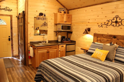 Cozy cabin interior with a bed, kitchenette, and wooden decor, featuring warm lighting and a welcoming door.