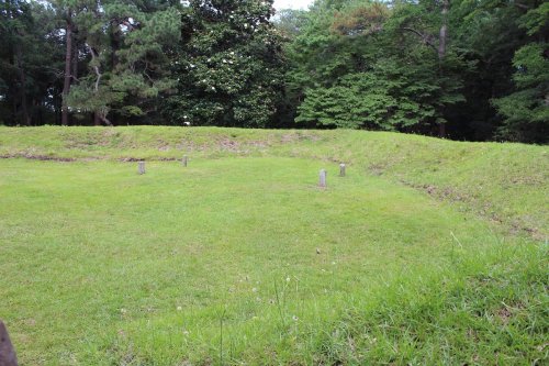 A grassy area with a circular earthwork and several stone markers, surrounded by trees in a natural setting.