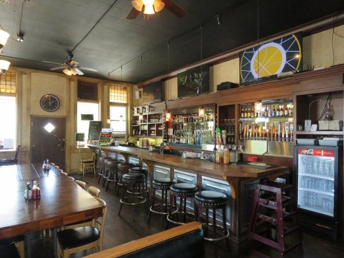A cozy bar interior with a wooden counter, stools, shelves of drinks, and a TV on the wall.