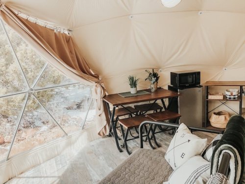 Cozy interior of a geodesic dome with a dining table, kitchenette, and large window showcasing nature outside.