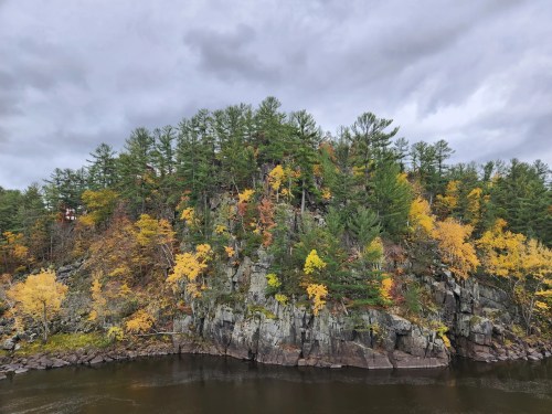 A rocky shoreline with vibrant autumn trees reflecting in calm water under a cloudy sky.