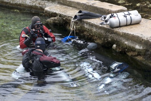 Two divers in wetsuits prepare to enter the water near scuba tanks on a rocky shore.