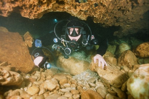 A diver explores an underwater cave, surrounded by rocks and pebbles, with a flashlight in hand.
