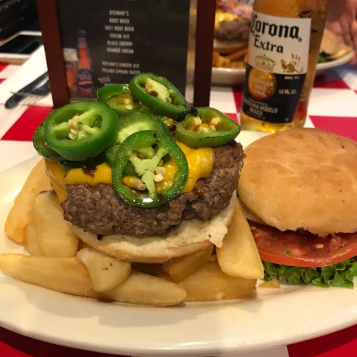 A juicy burger topped with jalapeños and cheese, served with fries and a side of a bun, with a beer in the background.