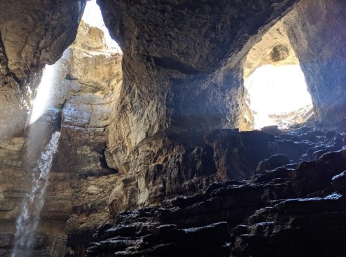 A view of a rocky cave with two openings, sunlight streaming in and water cascading down the stone walls.