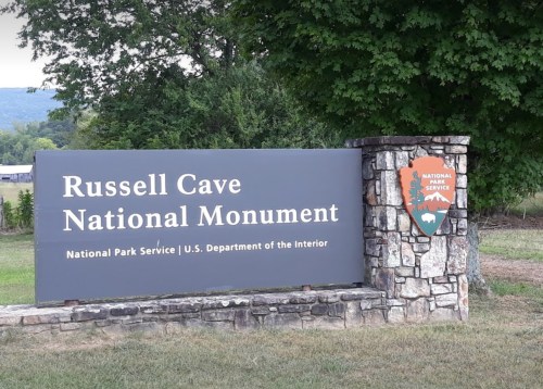 Sign for Russell Cave National Monument, featuring the National Park Service logo and surrounded by greenery.