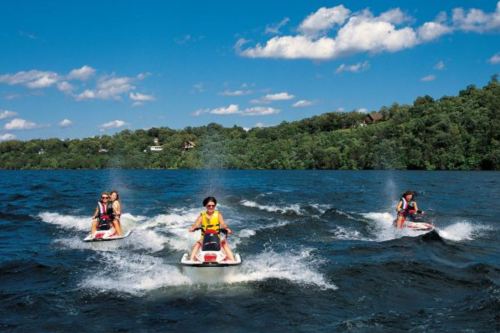 Three people riding jet skis on a lake, surrounded by green trees and a blue sky with fluffy clouds.