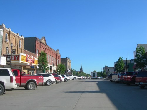 A sunny street scene featuring parked cars and historic buildings, with a theater marquee and a church steeple in the background.