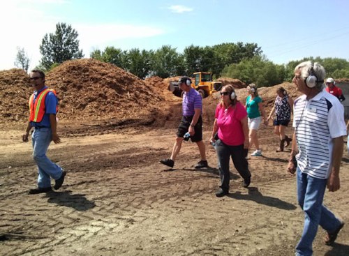 A group of people wearing ear protection walks through a wood chip yard, with piles of mulch in the background.