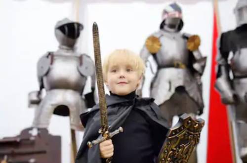 A young child in a black cape holds a sword and shield, standing in front of suits of armor.
