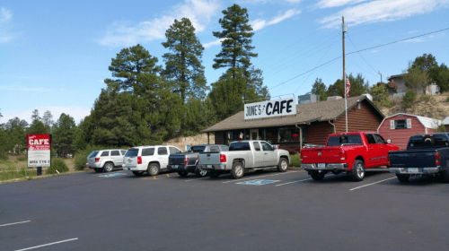 A rustic café building with a sign reading "June's Café," surrounded by trees and a parking lot filled with vehicles.