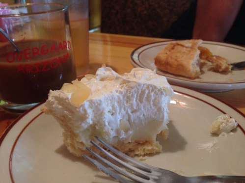 A slice of creamy dessert on a plate with a fork, alongside a glass of coffee and a partially eaten piece of cake.