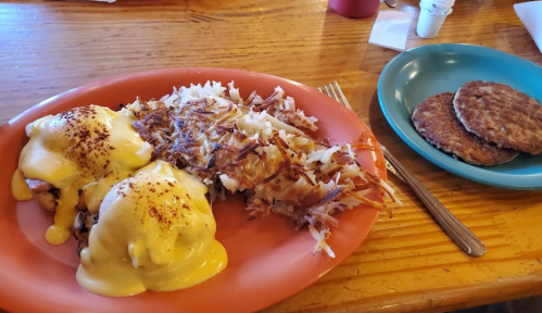 A plate of eggs Benedict with hash browns and a side of sausage patties on a wooden table.