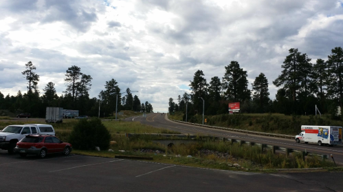 A view of a highway lined with trees under a cloudy sky, with parked cars in the foreground.