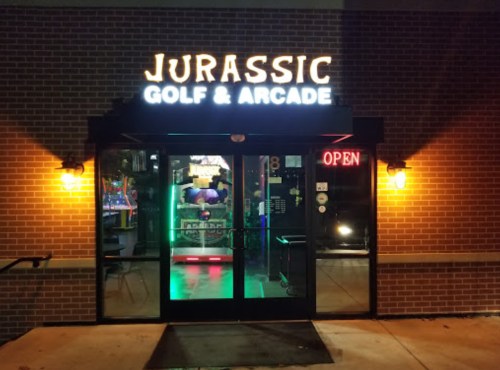 Entrance of Jurassic Golf & Arcade, featuring bright neon signage and an inviting open door at night.