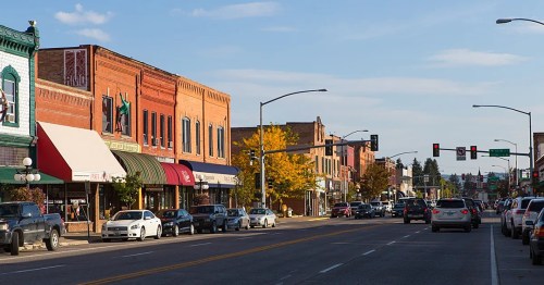 A bustling downtown street with brick buildings, shops, and cars under a clear blue sky.