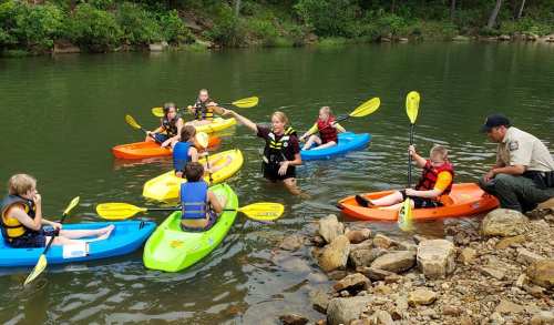 A group of children in colorful kayaks receives instruction from an adult by the water's edge.