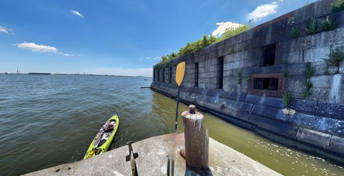A kayak is moored near a weathered stone wall by the water, under a clear blue sky with a few clouds.