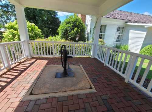 A vintage hand pump stands on a stone base, surrounded by a brick patio and white railing in a sunny garden setting.