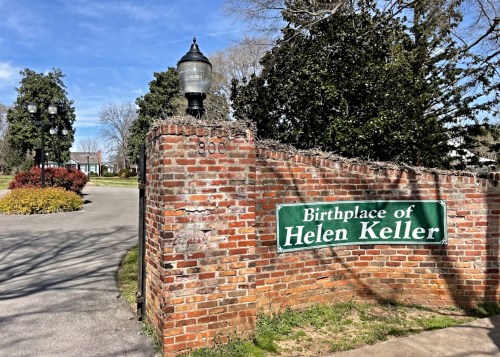 Sign marking the birthplace of Helen Keller, surrounded by greenery and a brick wall on a sunny day.