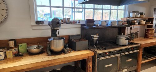 A rustic kitchen with wooden counters, a mixer, pots, and an old stove, illuminated by natural light from a window.
