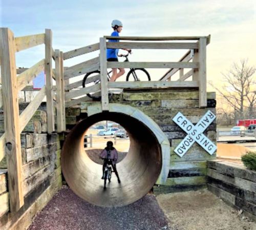 A cyclist rides over a wooden bridge while another cyclist emerges from a large tunnel below.