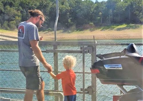 A man and a child stand by a lake, interacting near a boat, with trees in the background.