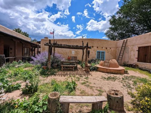 A rustic courtyard with a wooden pergola, plants, and a clay structure, surrounded by adobe buildings and a blue sky.