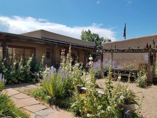 A charming adobe building surrounded by vibrant flowers and greenery under a blue sky with a flag in the background.
