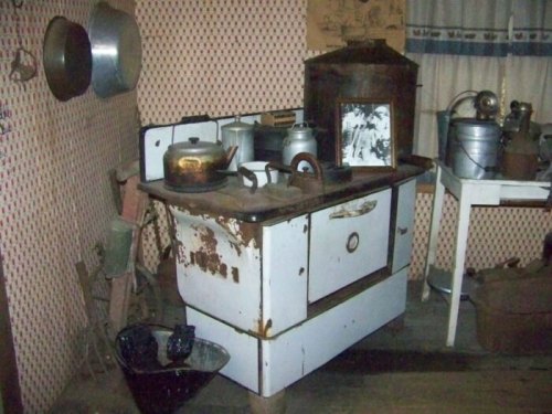 A vintage kitchen with an old stove, pots, and various utensils against a patterned wall.