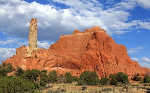 A tall rock formation rises from a red sandstone landscape under a blue sky with scattered clouds.