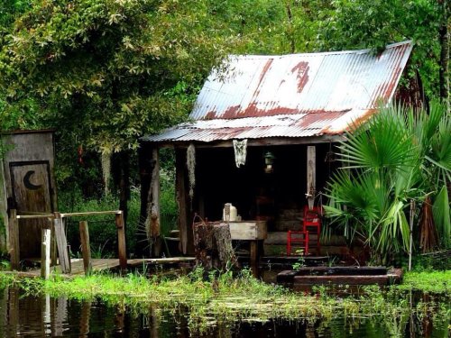 A rustic, weathered cabin with a metal roof, surrounded by greenery and water, featuring a porch and a red chair.