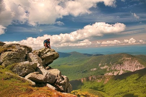 A hiker sits on a rocky ledge, overlooking a lush green valley and dramatic clouds in a bright blue sky.