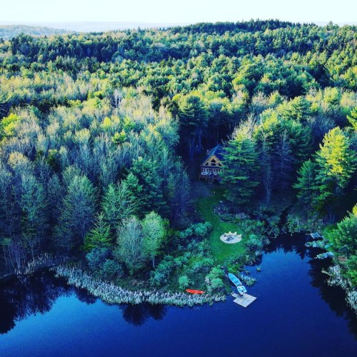 Aerial view of a serene forested area with a small house by a lake, surrounded by trees and colorful kayaks.