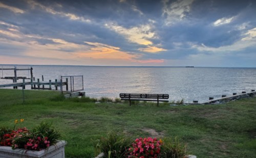A serene waterfront view at sunset, featuring a grassy area, flowers, a bench, and a distant pier.