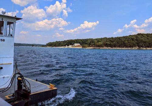 A view of a calm lake with a shoreline of trees and a building in the distance, seen from a boat.