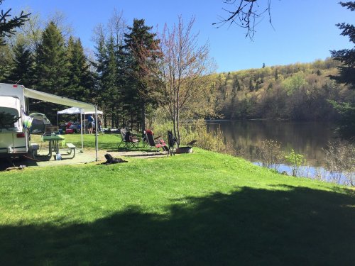 A serene lakeside scene with green grass, trees, a camper, and picnic setup under a clear blue sky.