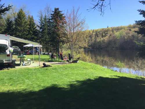 A serene lakeside scene with green grass, trees, a camper, and picnic setup under a clear blue sky.