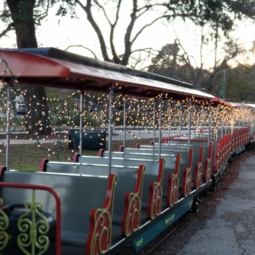 A festive train with empty seats, adorned with string lights, parked in a park during twilight.