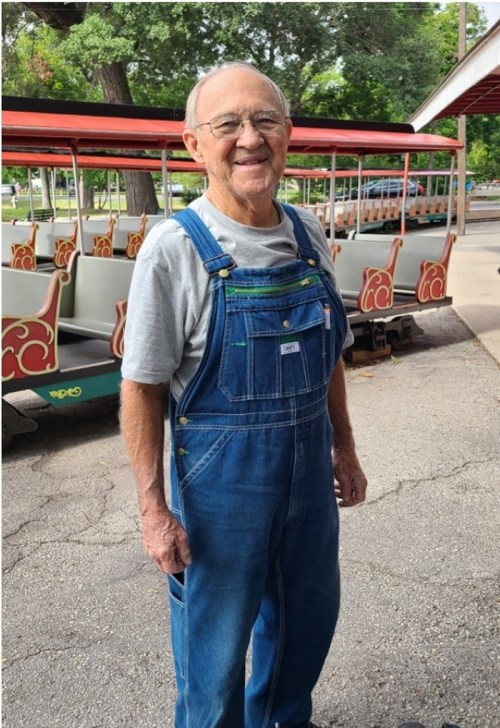 An elderly man in denim overalls and glasses smiles warmly in a park setting with a tram in the background.
