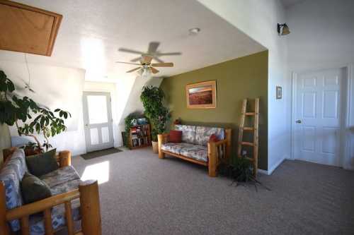 Cozy living room with wooden furniture, plants, and a green accent wall, featuring a ceiling fan and natural light.