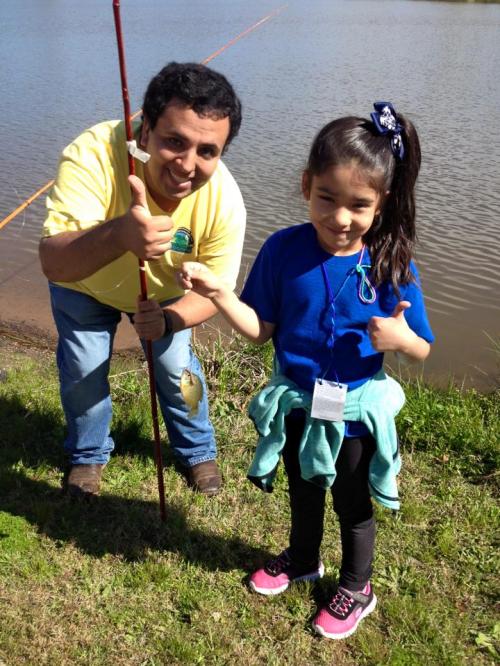 A man and a girl give thumbs up by a lake, holding a fishing rod with a fish on it. Both are smiling.