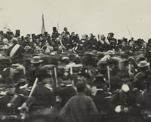 A large crowd of people in historical attire, gathered outdoors, with flags and hats, creating a lively atmosphere.