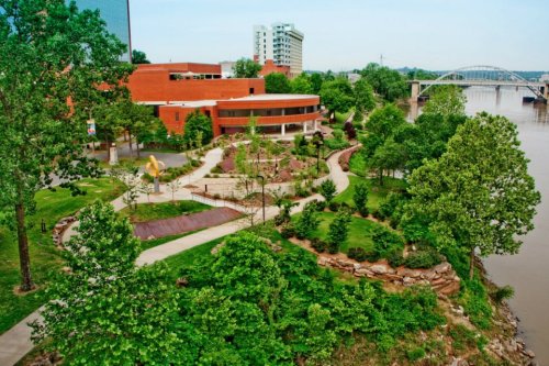 A scenic park with lush greenery, pathways, and a river view, featuring modern buildings in the background.