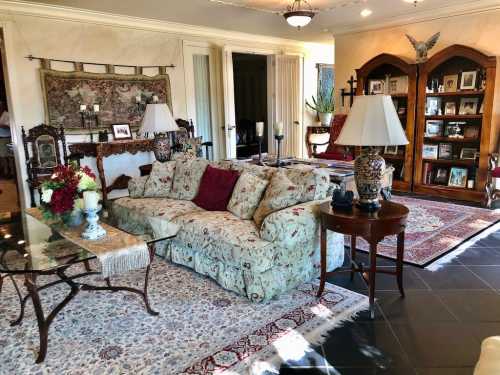 Cozy living room with floral sofa, glass coffee table, decorative lamps, and bookshelves filled with photos and decor.