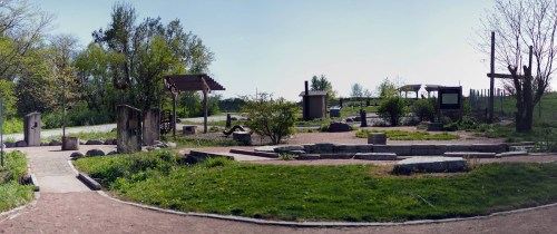 A panoramic view of a serene outdoor space with greenery, pathways, and wooden structures under a clear blue sky.