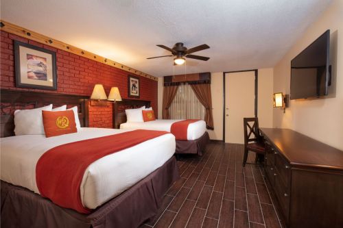 A cozy hotel room with two beds, wooden floors, a ceiling fan, and a TV, featuring warm red accents and artwork.