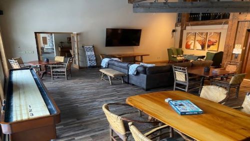 A spacious lounge area with wooden floors, a shuffleboard table, sofas, and dining tables, featuring warm lighting.