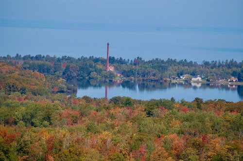 A scenic view of a lake surrounded by colorful autumn trees and a distant smokestack against a clear blue sky.