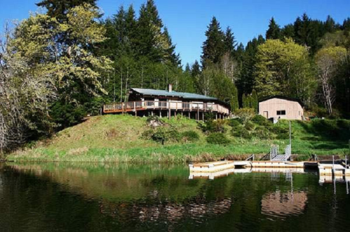 A serene lakeside view featuring a cabin and a dock surrounded by lush greenery and trees under a clear blue sky.